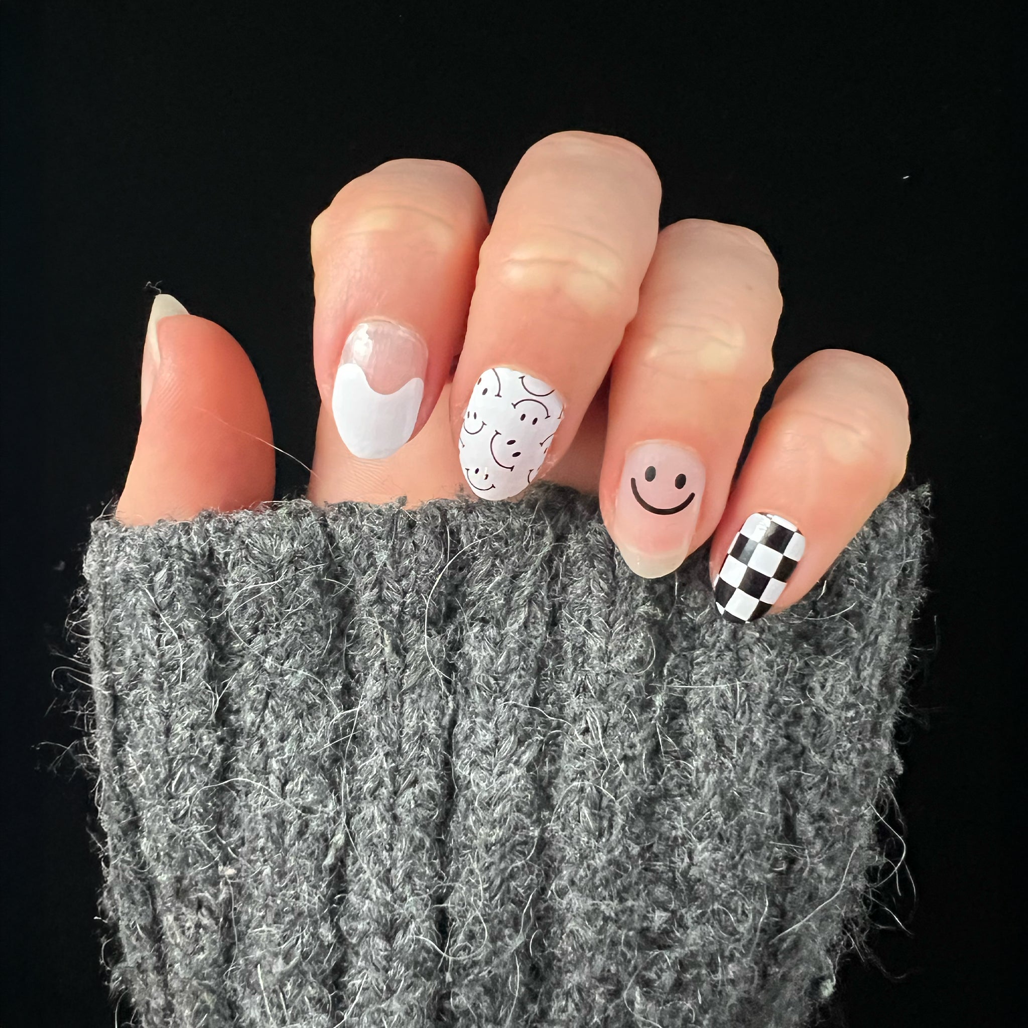 Hand met grijze wollige trui laat zwart-witte nagelstickers zien met smileys en blokjes