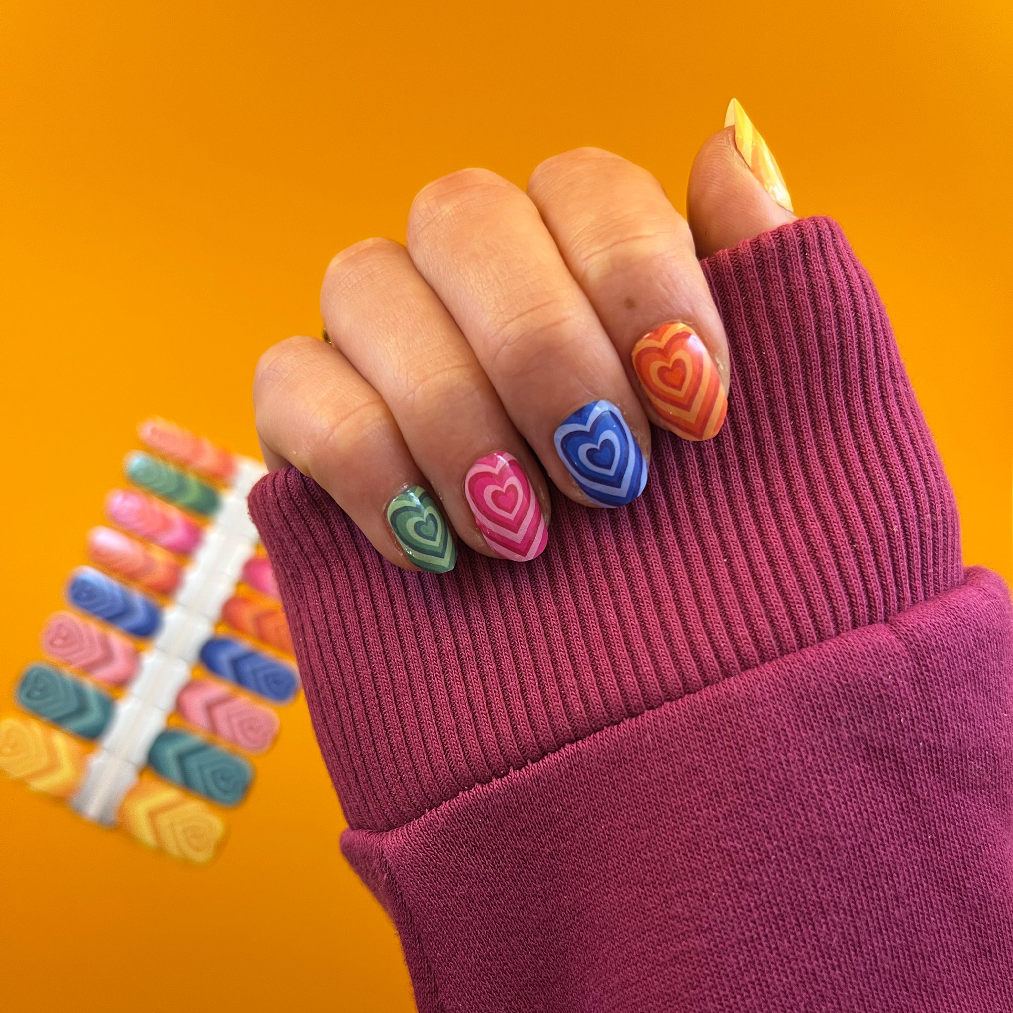 Nagels laten nagelstickers zien met kleurrijke hartjes op gele achtergrond en met donkerroze mouw