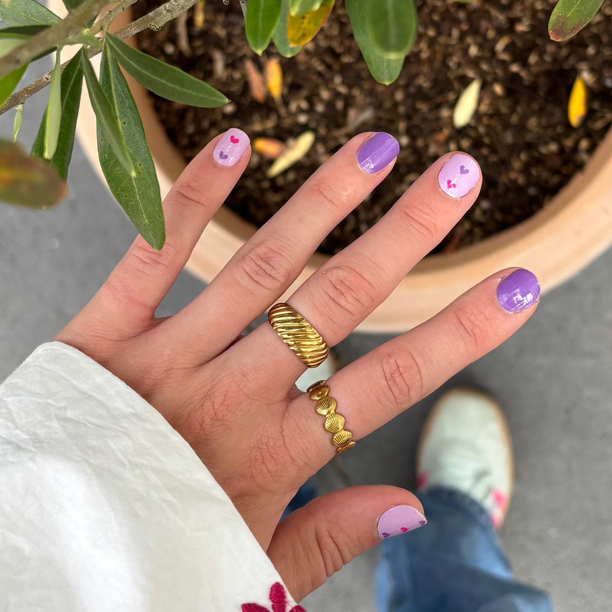 Hand met gouden ringen laat paarse nagelstickers met hartjes zien boven een plant