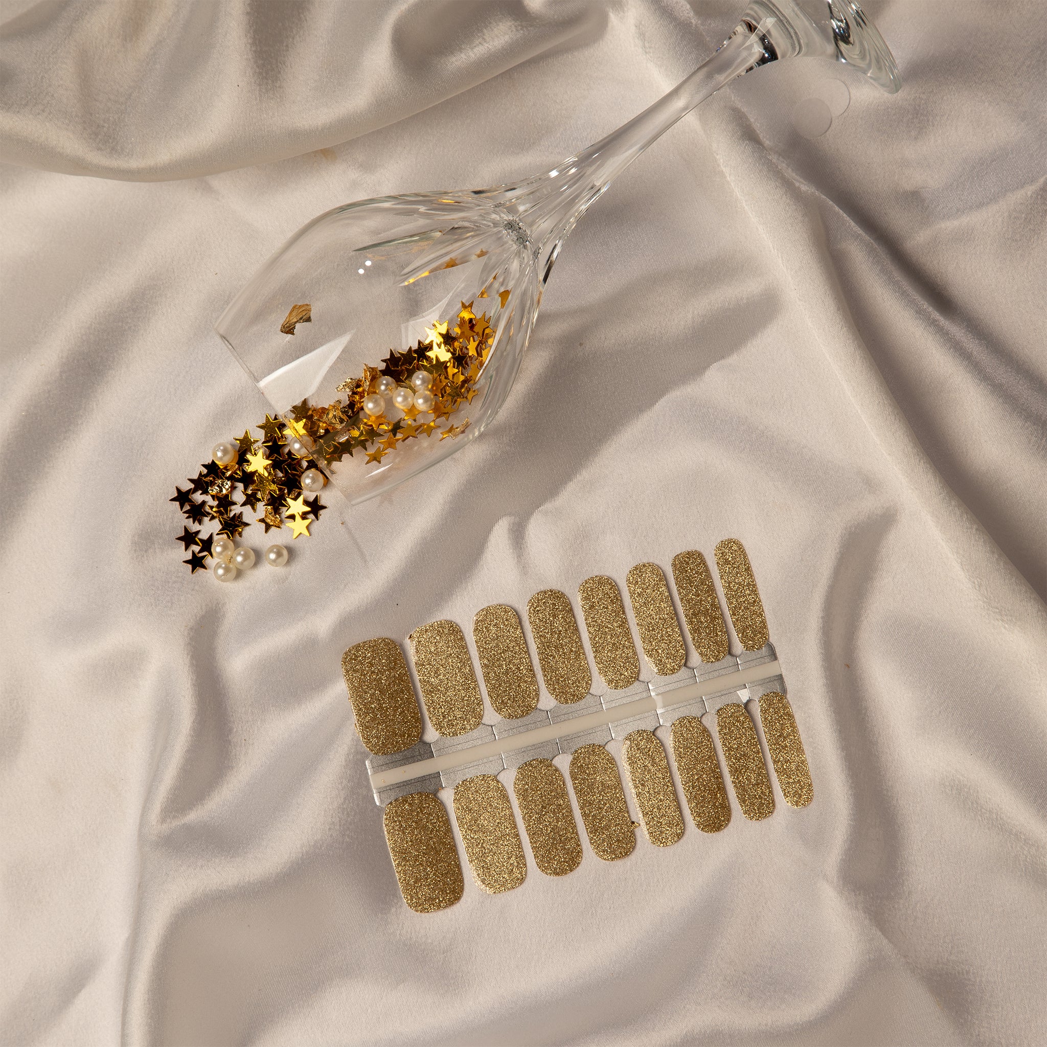 Gouden nagelstickers met omgevallen wijnglas me gouden glitters erin op wit satijnen doek