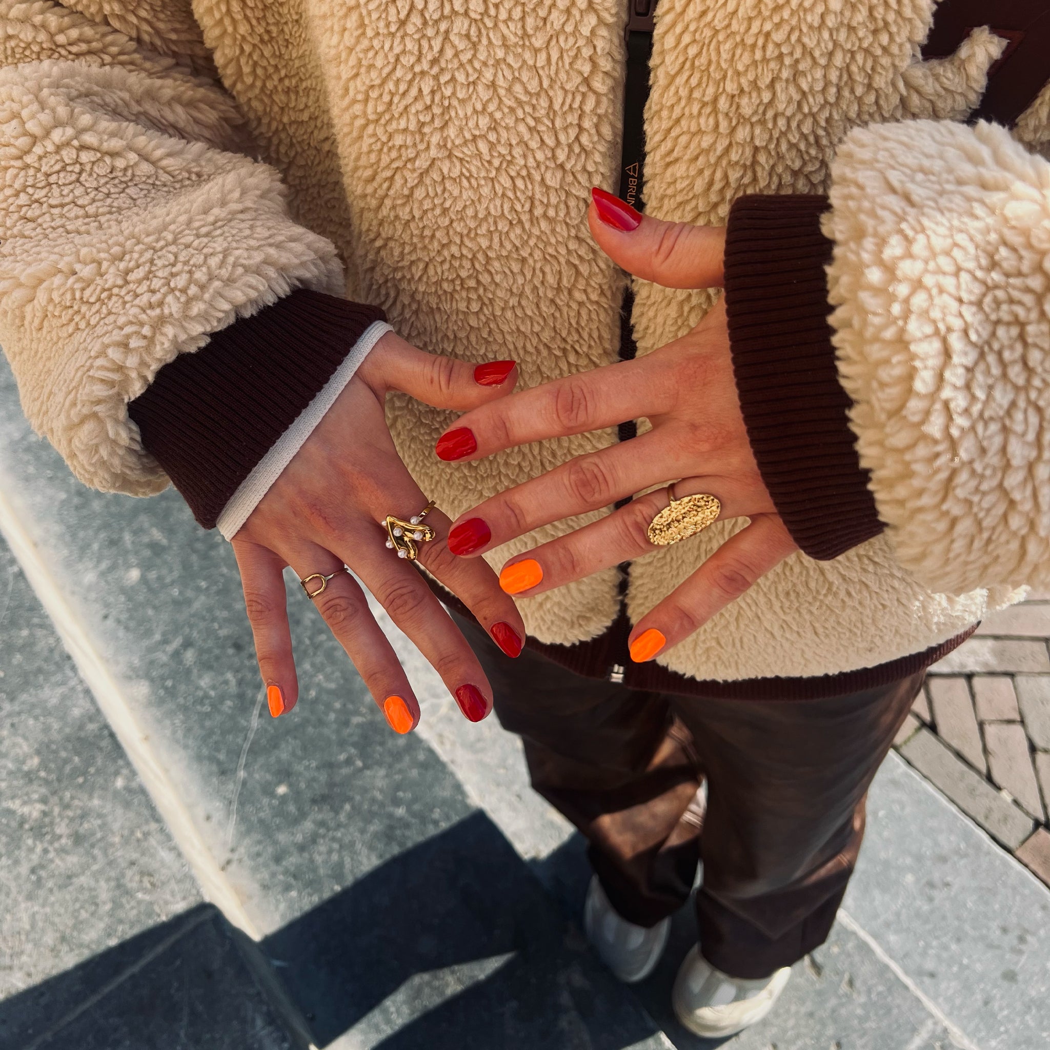 twee handen laten nagelstickers in rood en oranje zien met teddy vest en gouden ringen